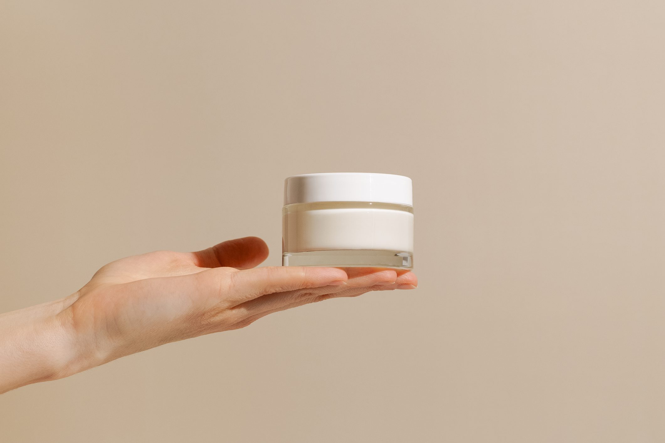 Hand holding a small white jar of cream against a beige background.